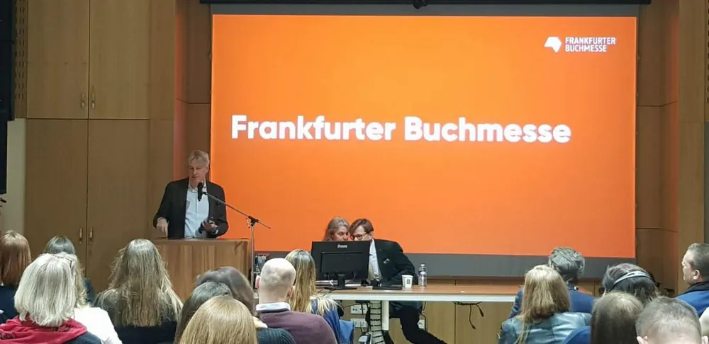 Seminář k českému hostování na frankfurtském knižním veletrhu. V popředí stojí řečník za pultíkem, zatímco za stolem sedí další dvě osoby sledující obrazovku počítače. V pozadí je velká oranžová obrazovka s nápisem "Frankfurter Buchmesse" (Frankfurtský knižní veletrh) a logem akce. Publikum sedí v hledišti před pódiem.